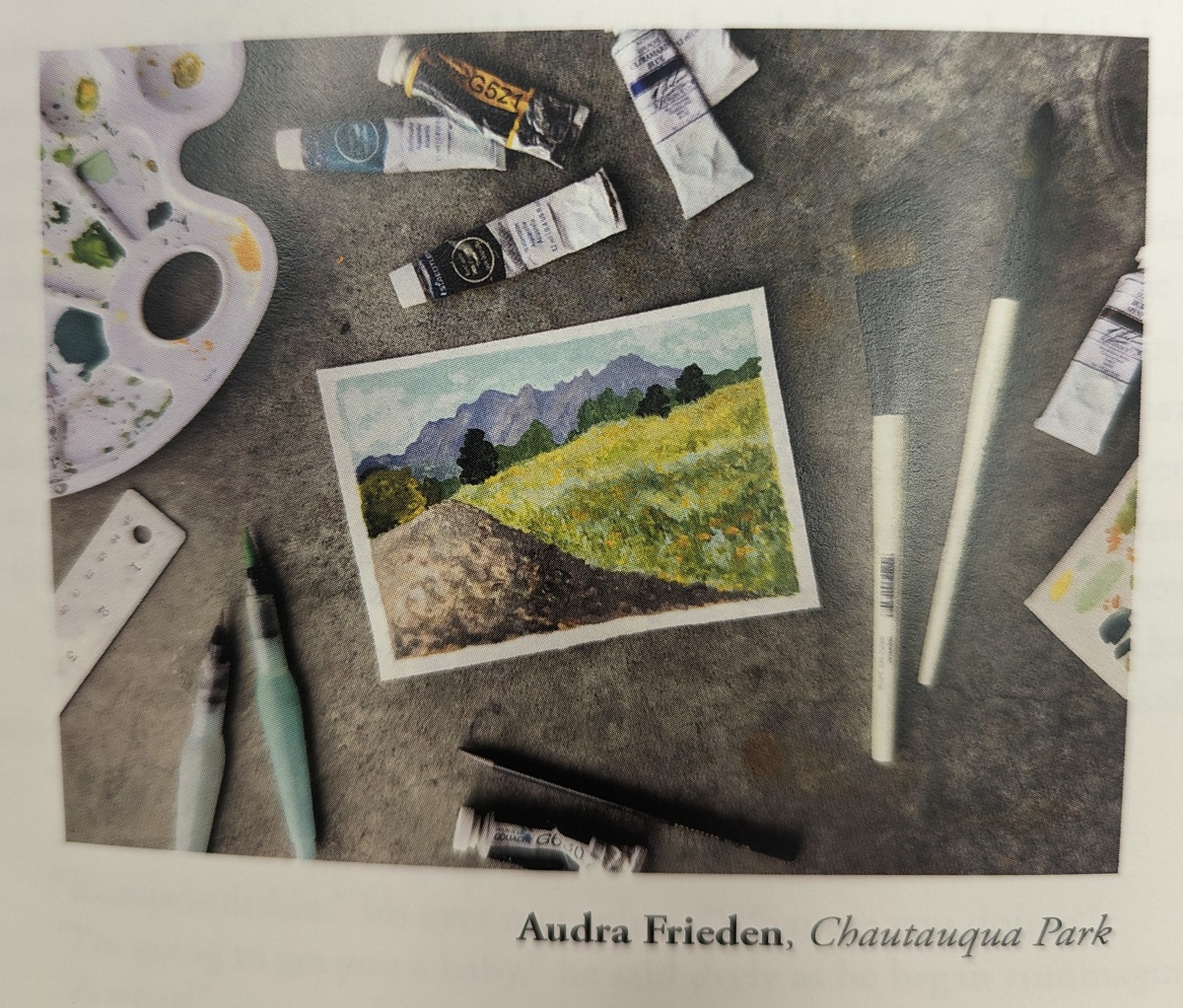 Image of a table filled with art supplies, with a small painting of the hillside in the center